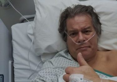 Man in hospital bed, giving a thump up with plaster and tube in this hand.