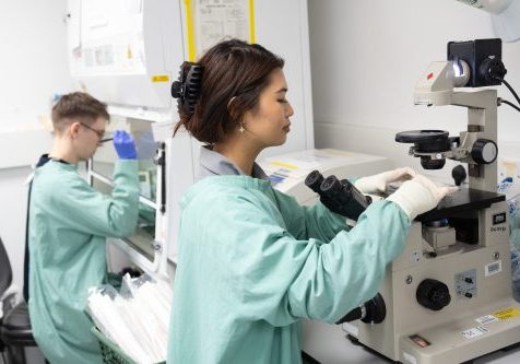 Male and female reserachers, standing side on, looking at microscopes. Both wear green lab coats.