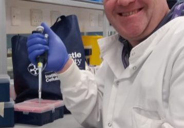 Professor Kevin Marchbank Male researcher, sat next to a lab bench. He is wearing a white lab coat and blue latex gloves and holding a pipette.