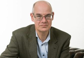 Professor Colin A Johnson Male sat on a brown leather sofa, draping his hands over the side. He is wearing a pale blue shirt and brown suit style jacket.