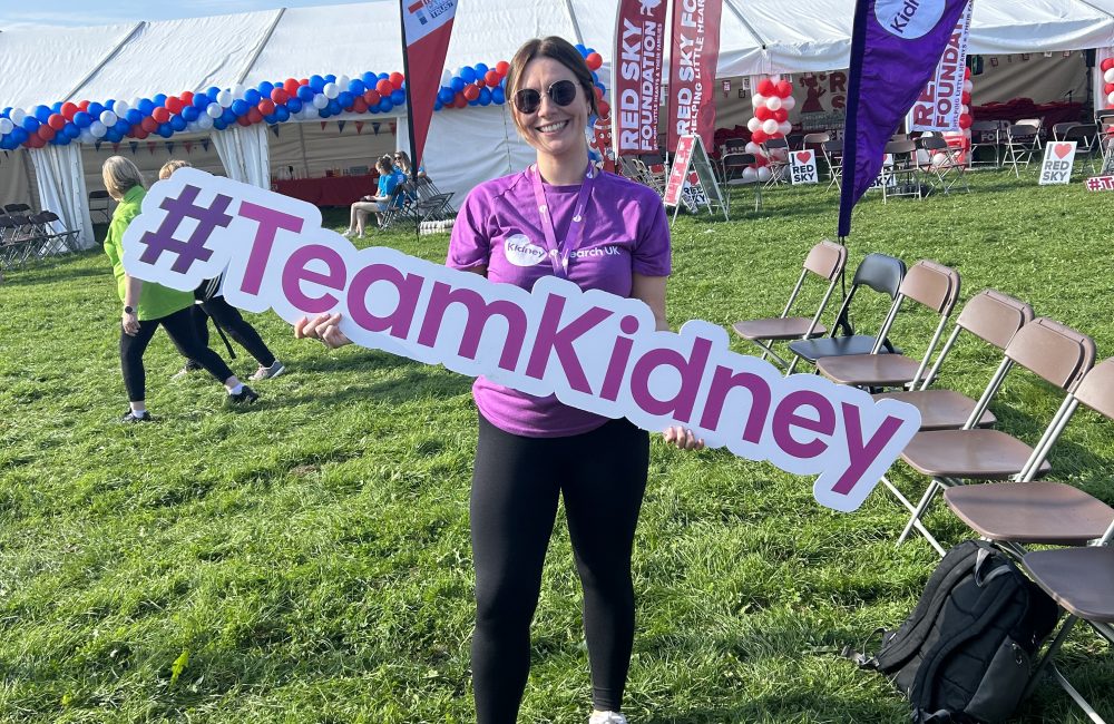 Female wearing black leggings, white trainers and a purple Kidney Research UK tshirt holding a foam #TeamKidney sign. A large white margee is behind her decorating in blue white and red balloons and some banners.