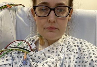 Woman in hospital gown sitting on chair taking a selfie