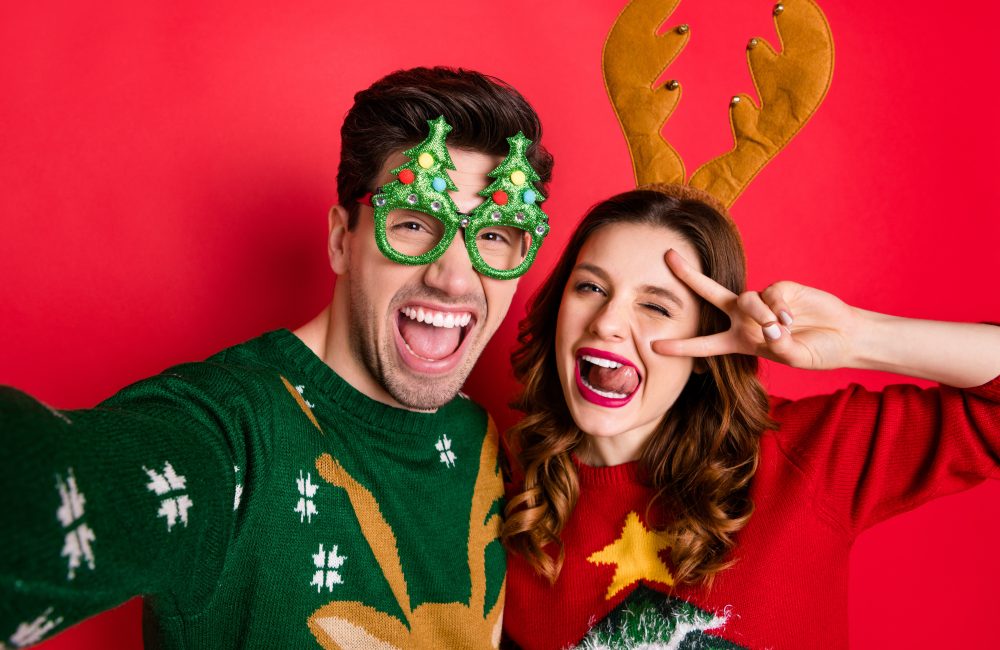 Photo of crazy couple making selfies sticking tongues winking eyes v-signing, wear funky ugly ornaments jumpers isolated red color background