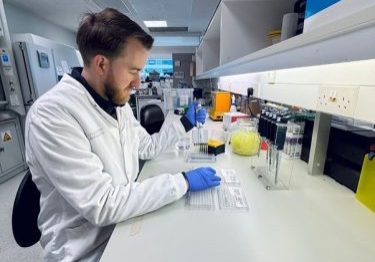 A male researcher, sat at a lab bench, wearing a white lab coat and blue glove, holding a pipette