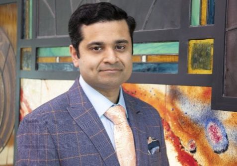 Krishna wearing blue checked suit with pink tie, smiling got the camera