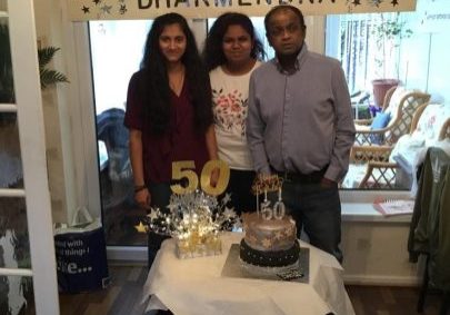 two females and male, standing at a table with a birthday cake. Banner hanging behind them saying Happy 50th Birthday