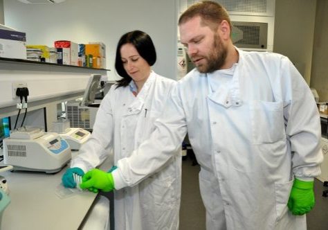 Dr. Oni and Dr. Chetwynd in the research lab wearing white coats and green latex gloves and looking at samples in a test tube