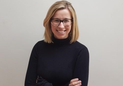 A lady with blonde bobbed hair, black glasses and a black jumper with her arms folded, smiling to the camera.