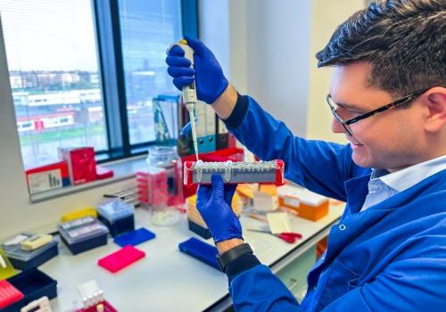 Man standing side on at a research lab bench holding research instrument. He's wearing a blue lab coat and blue gloves.