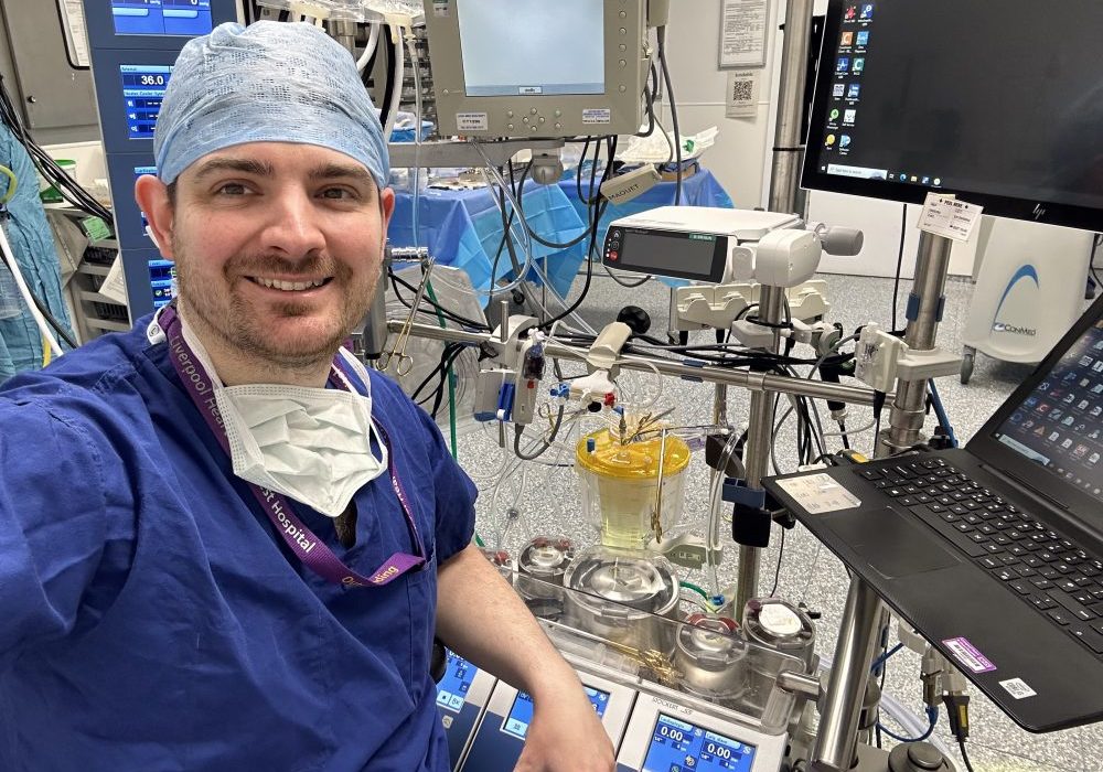 Mr James Bennett A male, dressed in blue hospital scrubs, in a operating room surrounded by equipment and a computer next to him,