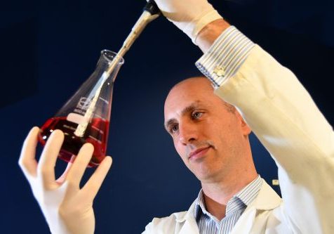 A man in white lab coat, surgical gloves holding a glass flask with liquid in