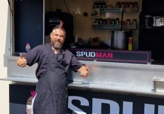 Man, with pink Mohican and wearing a black apron. He is standing in from of his food van giving a double thumbs up