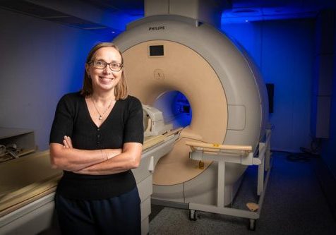 Female, wearing glasses with her arms folded, standing in front of an MRI machine