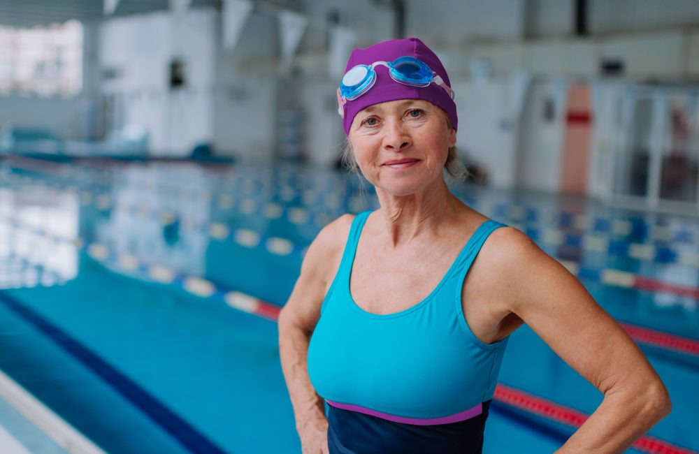 Female swimming at the pool, wearing a blus costume, purple swim cap and goggles on her head.