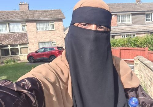 Close up of an Asian female, wearing a tan colour and black burka. Houses in the background