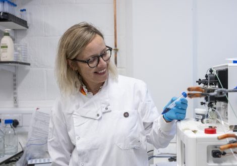 Female with blonde hair, in a white lab coat and round black glasses in a research lab