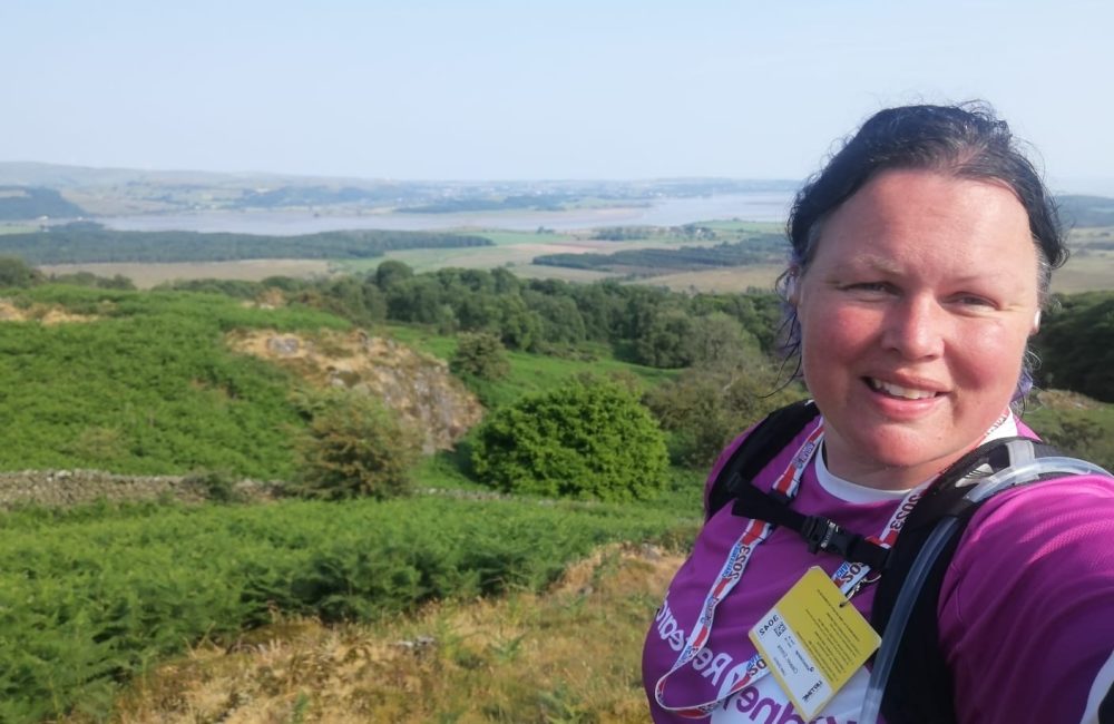 Feamle runner, wearing \Kidney Research UK t-shirt and a hydration pack, taking a selfie with the green landscape behind.