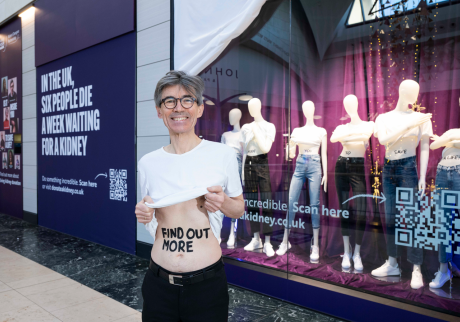 Middle-aged man with glasses stands in front of shop window which displays six mannequins with text on their stomachs spelling out 'Is it inside you to save a life?'. The man is lifting up his white t-shirt with the words 'find out more' on his stomach.
