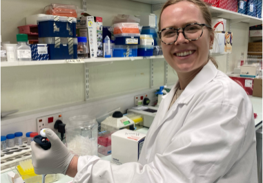 Laura, standing at her lab bench, wearing a white lab coat.