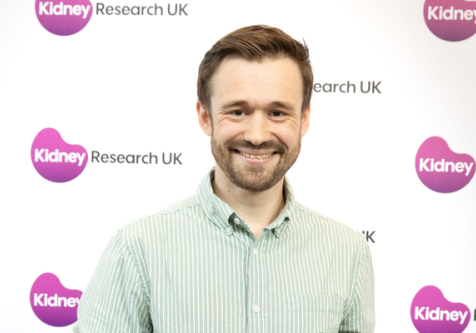 Sam smiling for the camera, he is wearing an green stripe collared shirt and Kidney Research UK logos are behind him